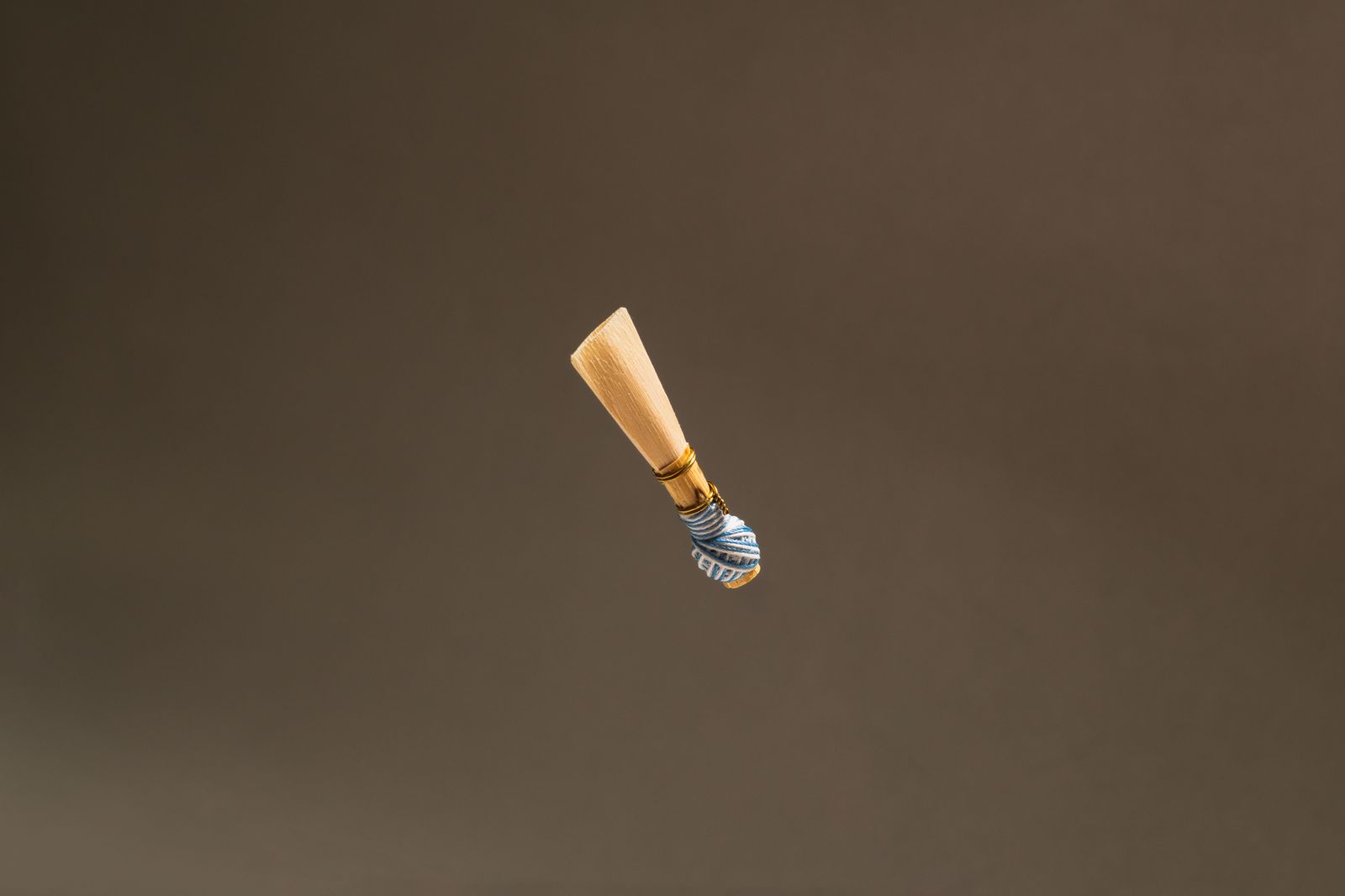 Een riet voor een blaasinstrument, omwikkeld met blauw garen, zwevend tegen een egale bruine achtergrond